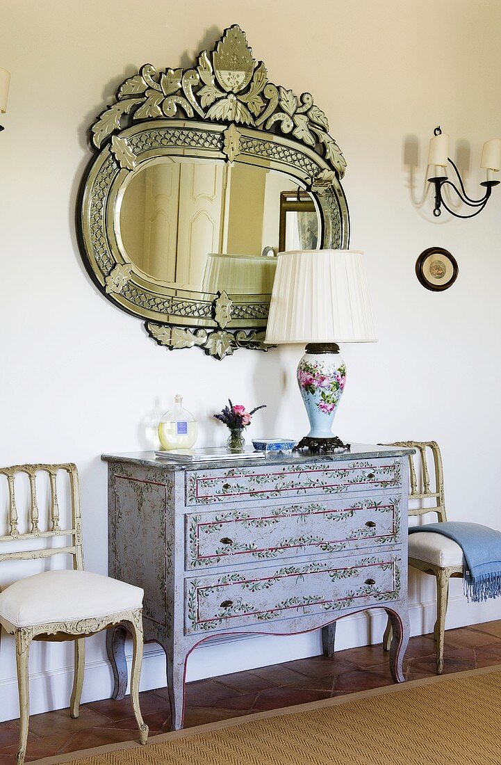 A mirror hanging above an antique chest of drawers with Rococo chairs standing against the wall in a hallway