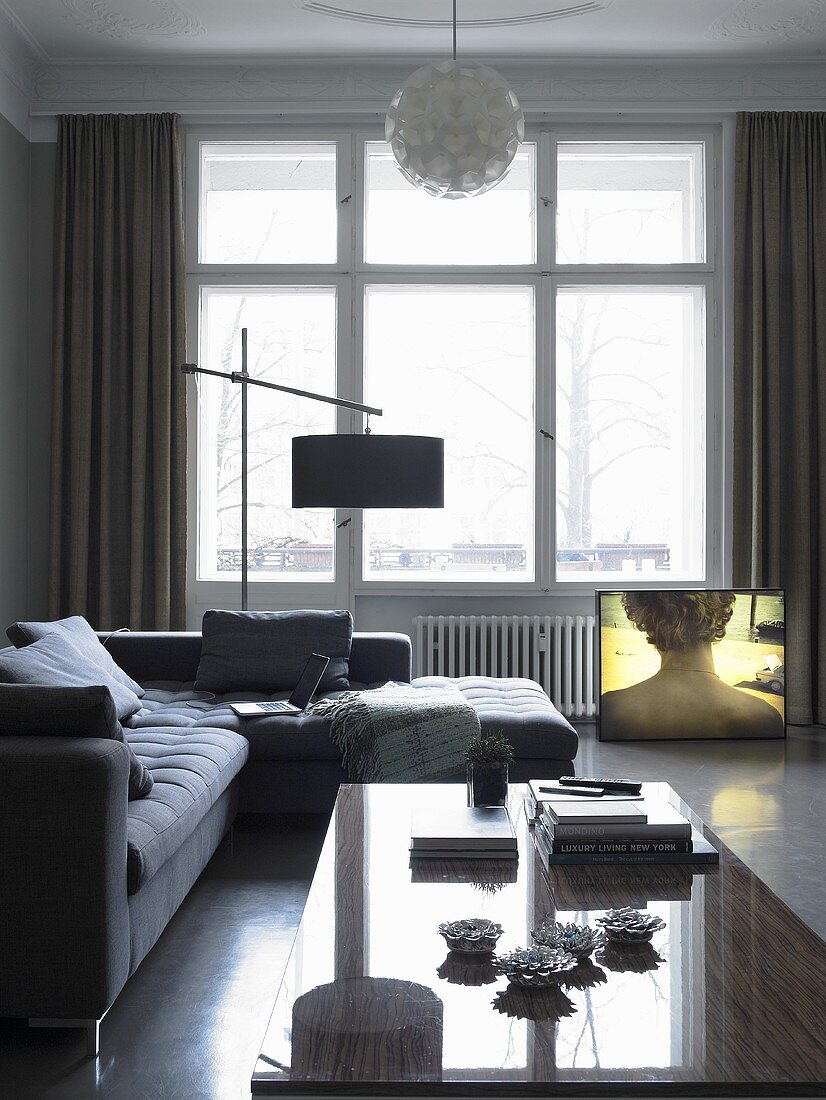 An upholstered corner sofa and a reflective coffee table in a living room with a floor lamp in front of a window
