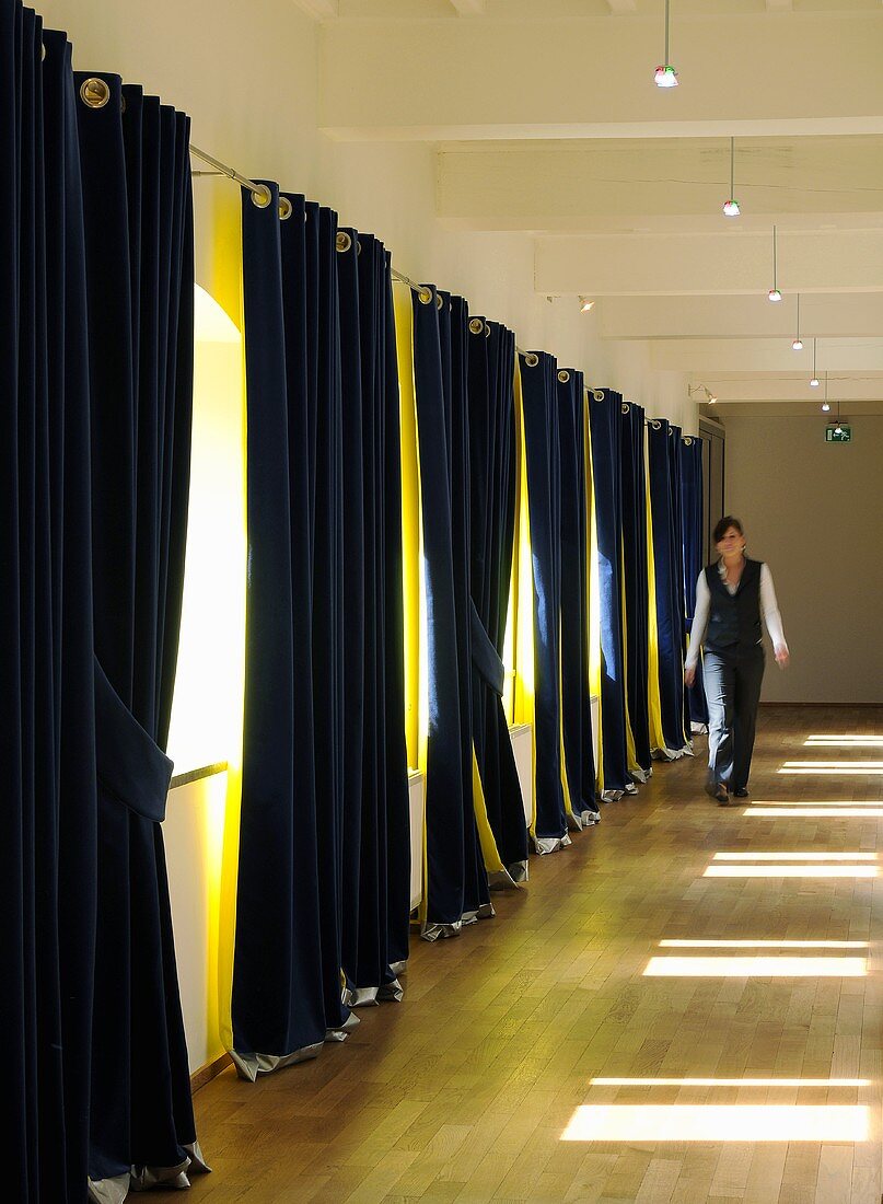 A woman in a corridor with half-closed, two-tone curtains hanging at the windows