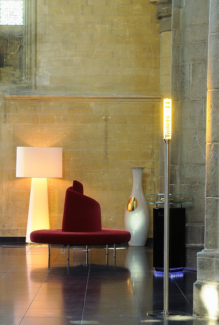 A designer seating area in a renovated church - a red circular bench and a floor lamp against a natural stone wall