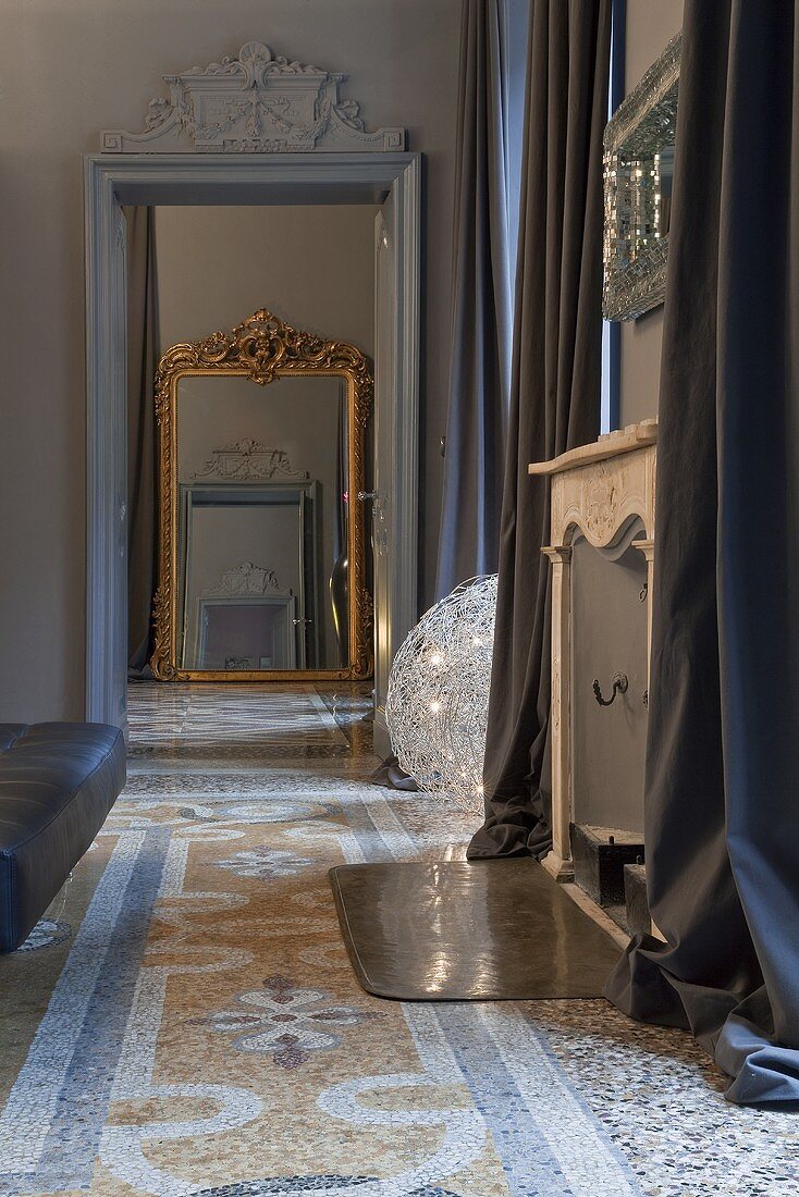 An anteroom with a view through a door onto a wall mirror with a gold frame and a patterned terrazzo floor
