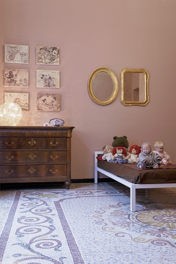 A child's bedroom - an antique chest of drawers and a collection of dolls on the bed in front of a pink wall and a patterned terrazzo floor