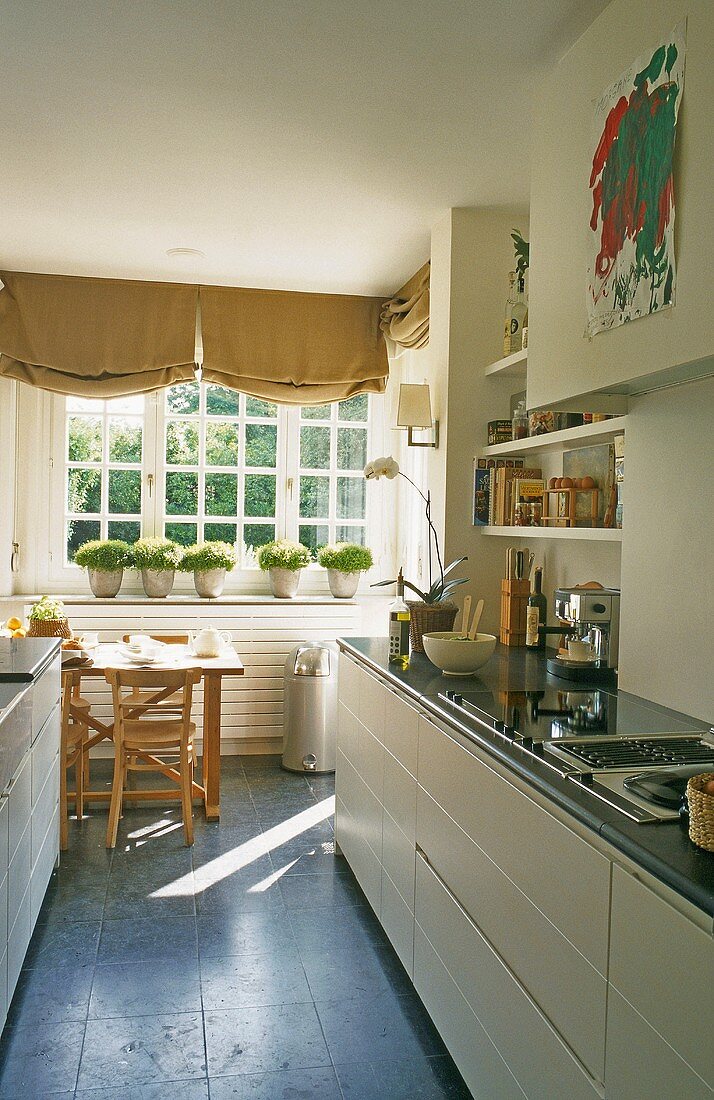 A narrow kitchen - a counter with white cupboards and a dining area with pots of herbs on the window sill