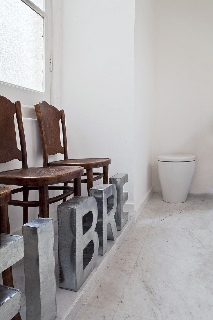 Künstlerische Rauminstallation - Metallbuchstaben vor antiken Stühle und weißem Behälter