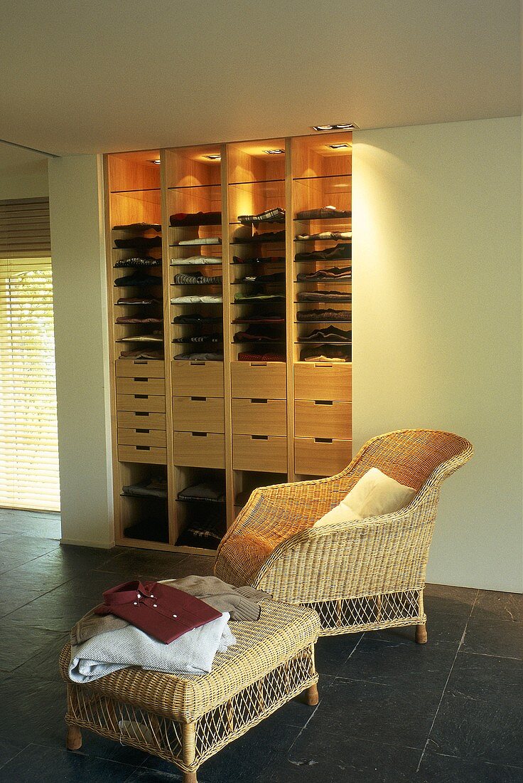 A dressing room with a wicker chair and a side table in front of a built-in, illuminated cupboard