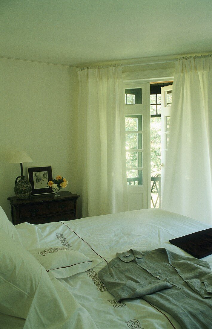 Clothes on a bed and white curtains in front of a balcony door
