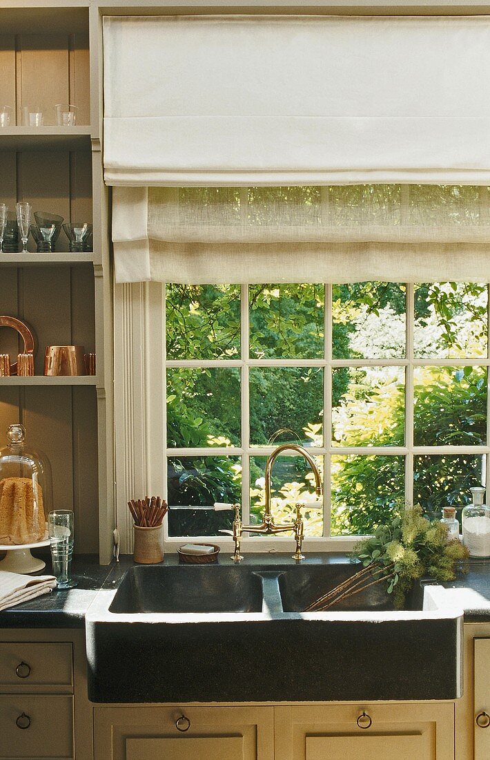 A kitchen in a country house with a view of the garden - a stone sink and brass taps in front of a transom window with a blind