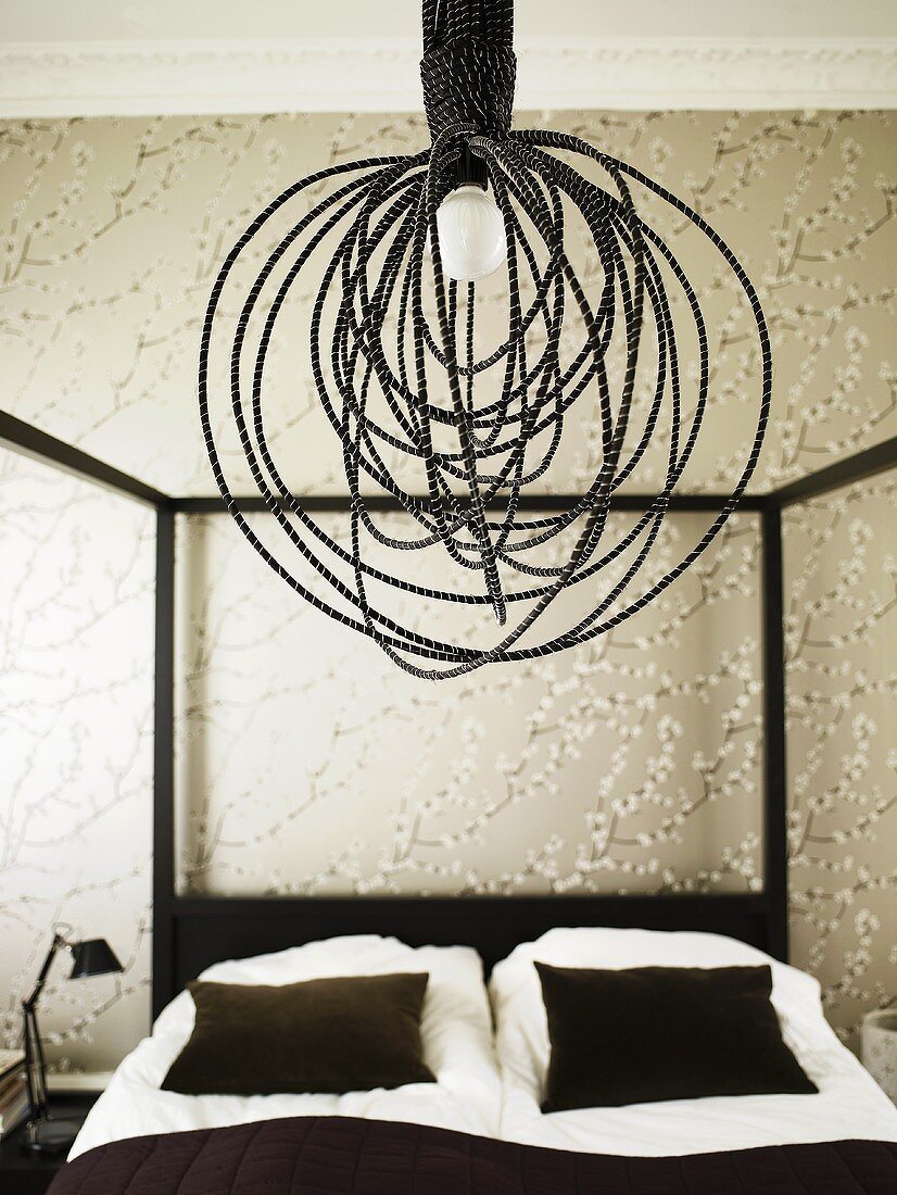A hanging lamp wrapped with black draw cords over a canopied bed in front of wall with wall paper