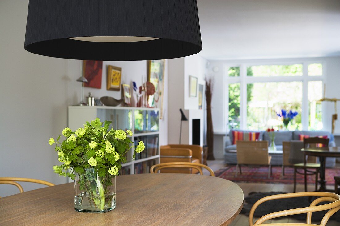 Black lampshade above flowers in a vase on a wooden dining table and an open plan living room