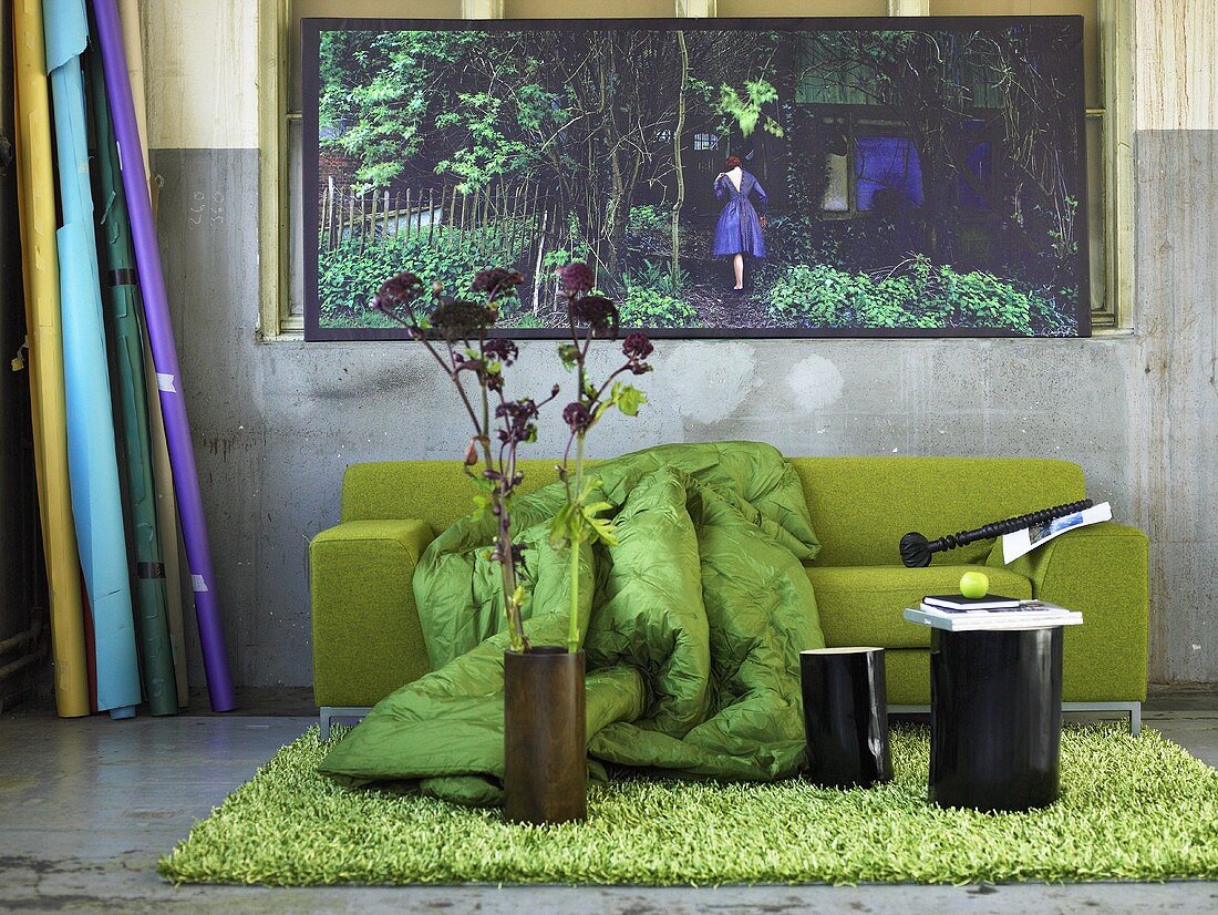 Blumen in Vase und schwarze Hocker auf grünem Flokatiteppich vor grünem Sofa mit Plaid