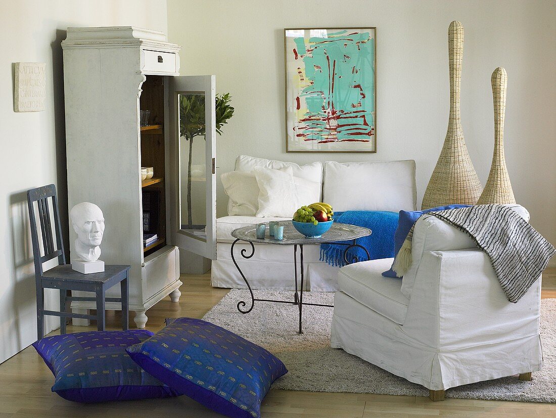 weiße Sofagarnitur mit blauen Bodenkissen und Vitrine mit offener Tür im Landhausstil