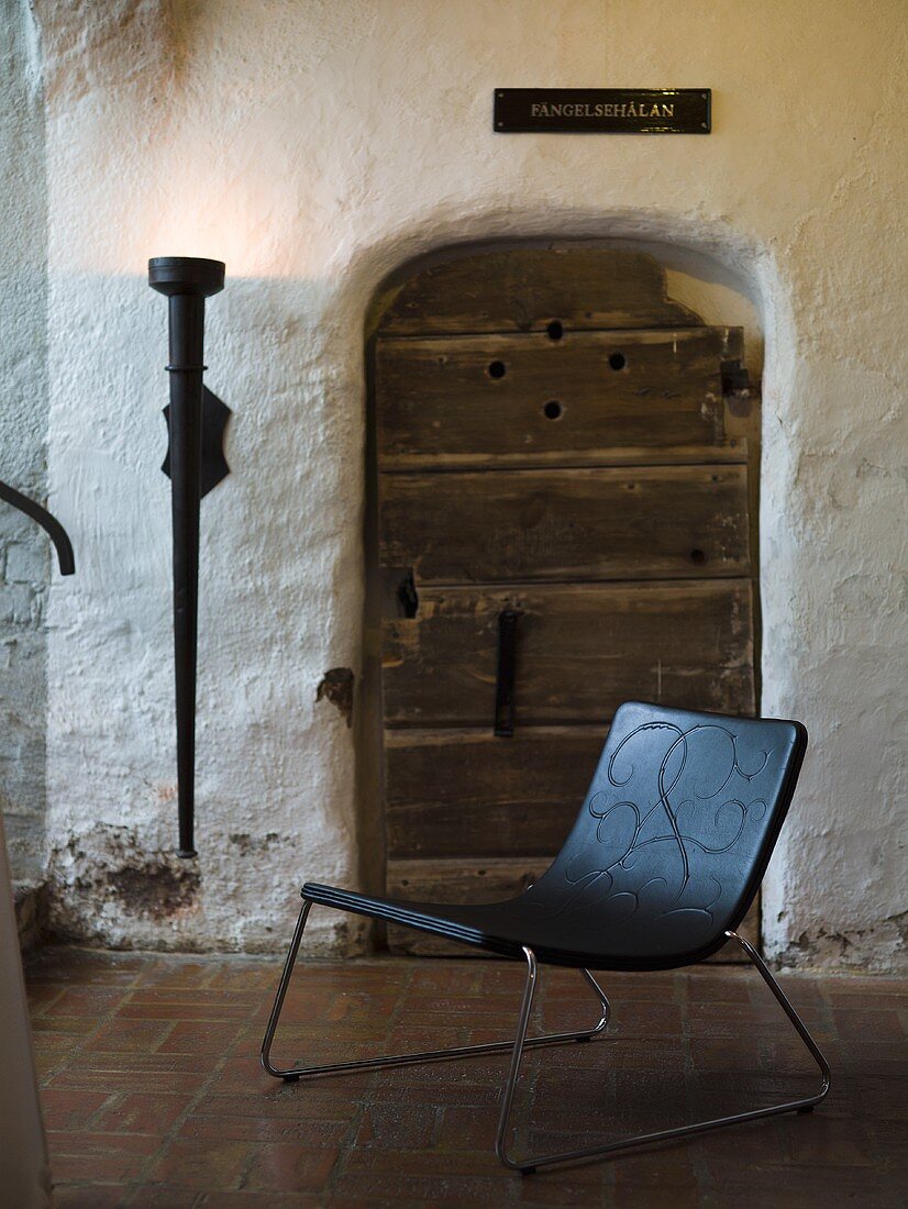 Schwarzer Lederstuhl in Schalenform vor alter Holztür und Wandbeleuchtung eines Schlosses