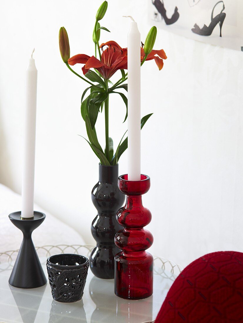Red and black candlesticks next to a flower vase with red lilies on a glass surface