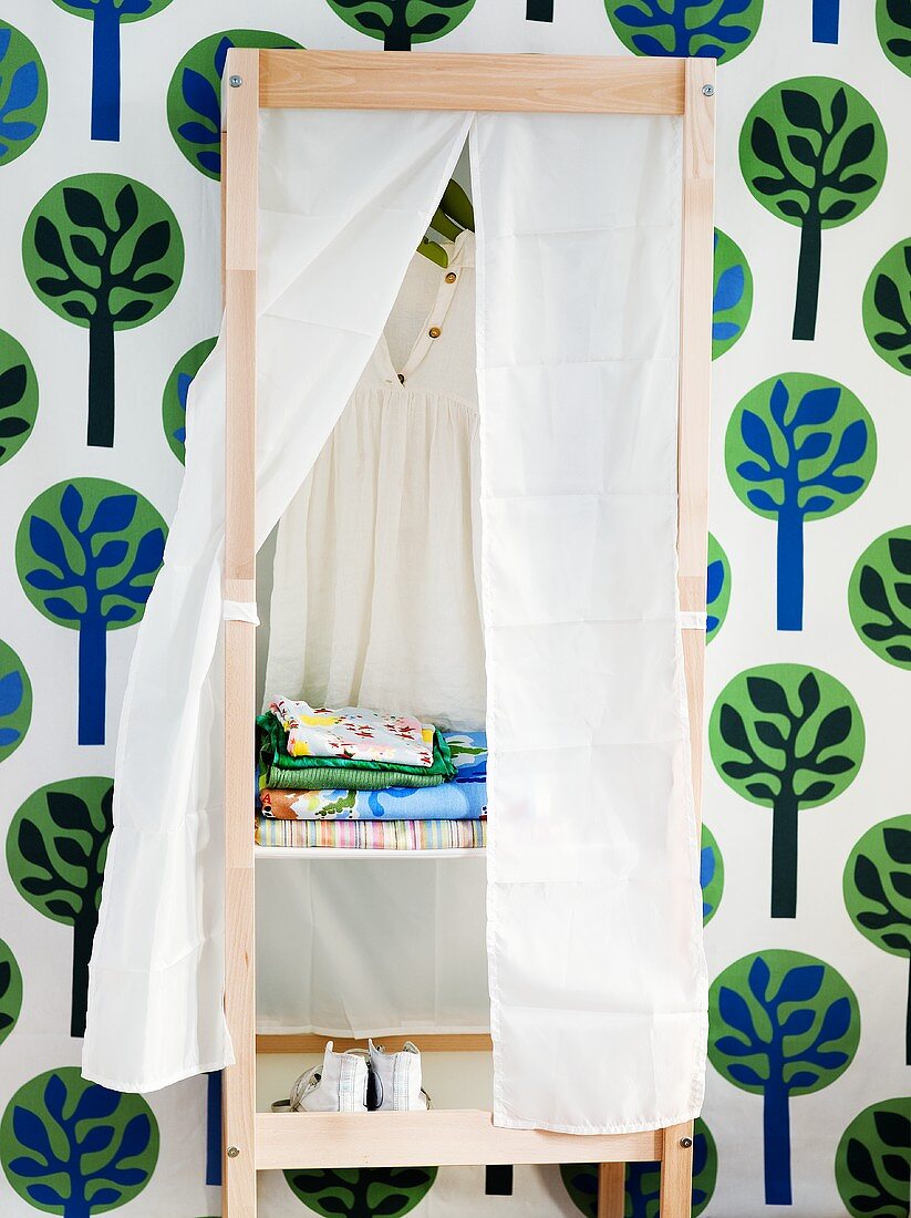 Holzregal mit geöffnetem weissen Vorhang und Blick auf Kleidung vor Tapete mit Blumenmuster