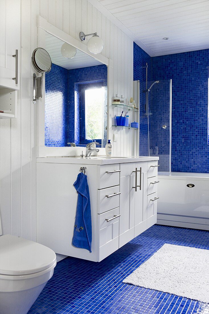 Waschtisch mit Spiegel im Bad - weiße Holzverkleidung an Wand und Decke und blaue Mosaikfliesen auf Boden und Wand um Badewanne