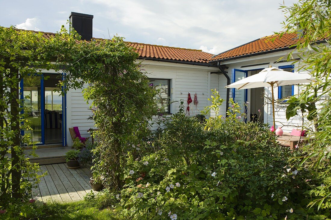 Garten mit beranktem Torbogen vor Terrasse eines weissen Holzhauses