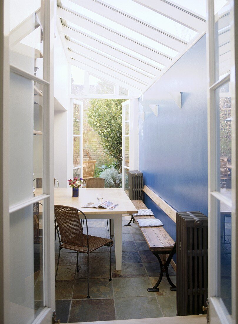 Blick durch eine offene Tür in Wintergarten mit Tisch und Sitzbank vor blauer