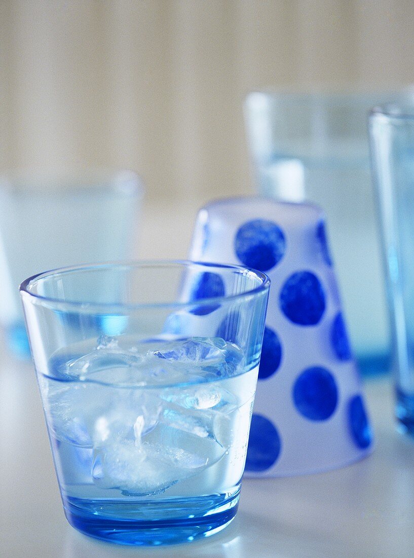 Blau getöntes Glas mit Wasser und Eiswürfeln vor Glas mit blauen Punkten