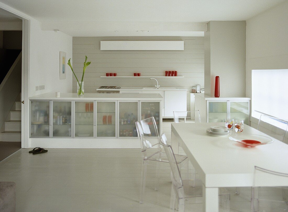 Modern, white open plan kitchen diner with glass fronted cupboard units and table with perspex chairs.