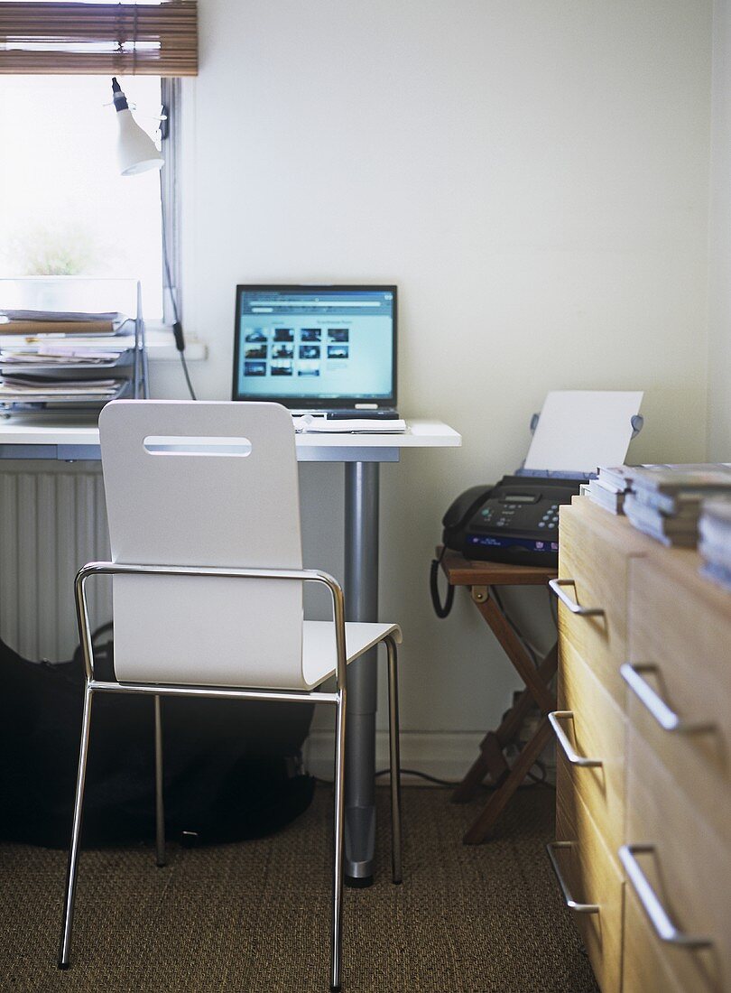 A modern home office with desk, chair, laptop computer, trays, drawers, fax , lamp, radiator,