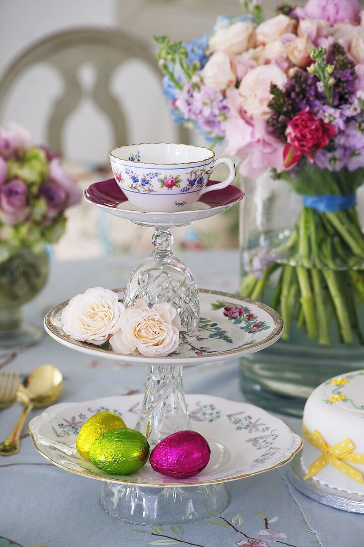 Etagere mit Teetasse, Rosenblüten und Schokoladeneiern vor Blumenstrauss
