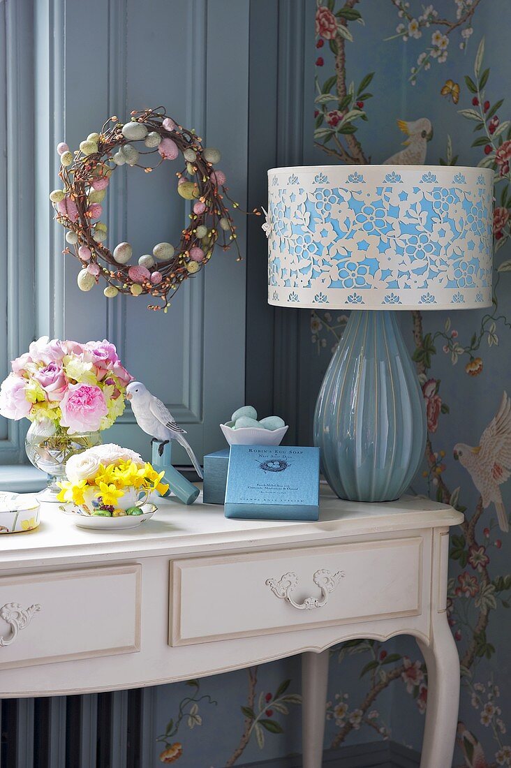 A table lamp with a patterned shade and flowers on a country house style wall table