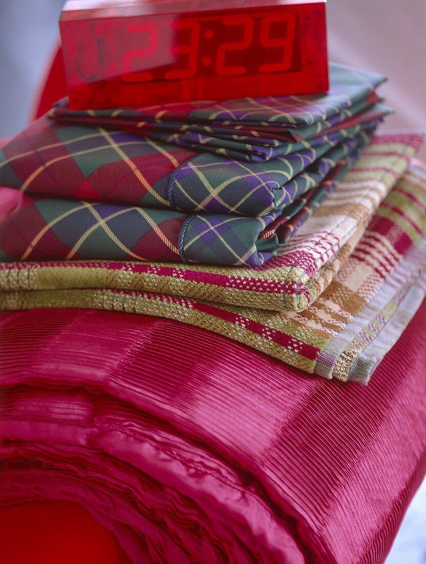 A detail of folded linens, pink textured throw, check towels, cube alarm clock,