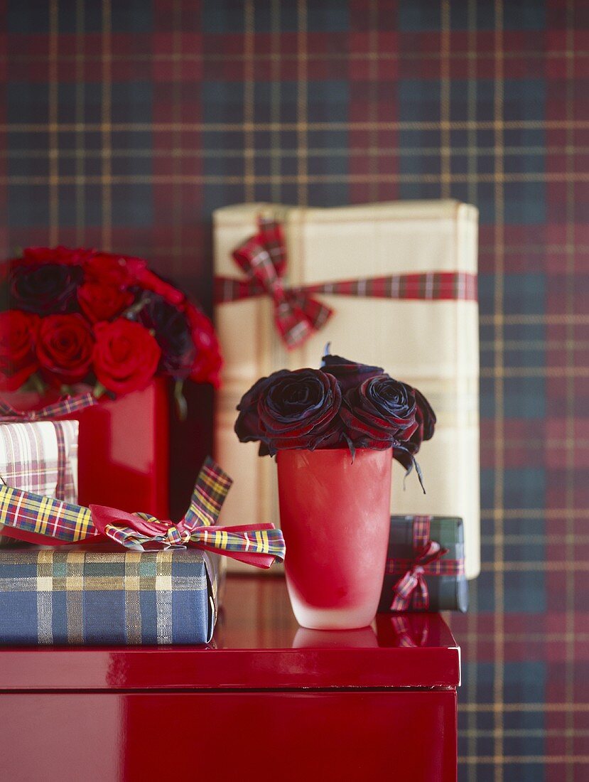 Rote Vase mit Rosen neben Geschenkpaketen vor karierter Tapete an Wand