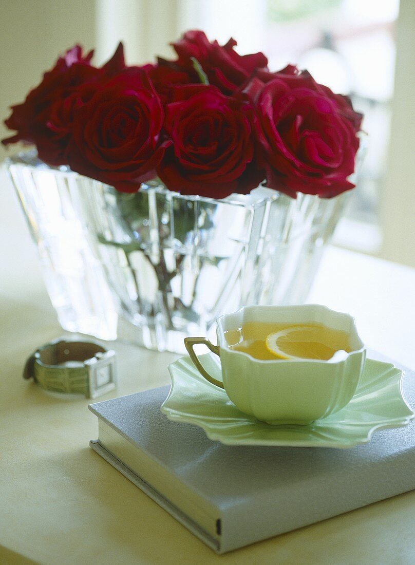 A detail of a cup of lemon tea resting on a book, red roses in a glass vase