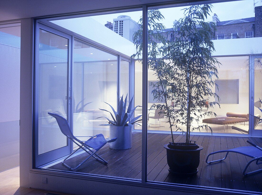 Blick durch raumhohe Fensterfront auf Terrasse mit modernen Gartenmöbeln und Topfpflanze