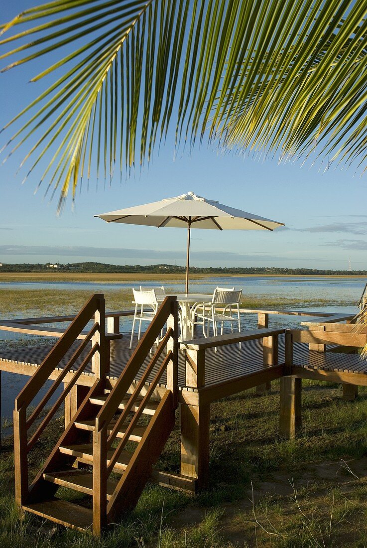 Ein erhöhtes Holzdeck mit Tisch, Stühlen und Sonnenschirm unter einer Palme am Meer