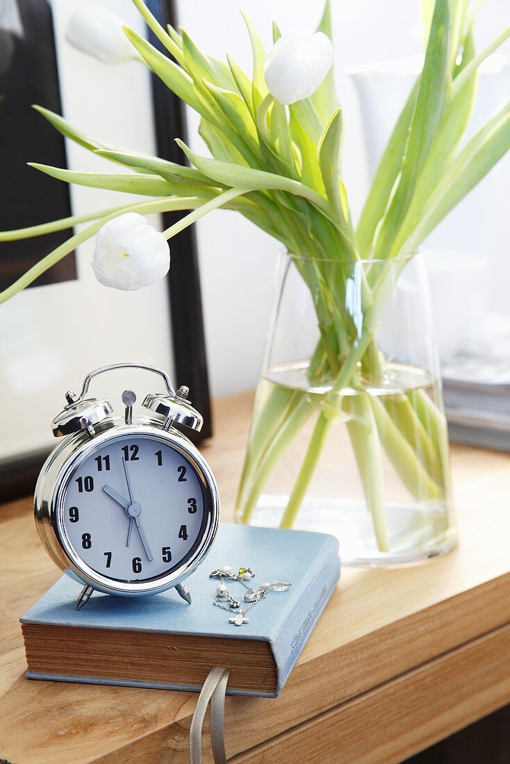 An alarm clock, a book and white tulips of a bedside table