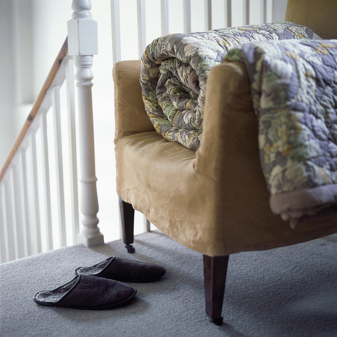 Ein Sessel mit zusammengerollter Steppdecke und Hausschuhe an der Treppe eines Flurs