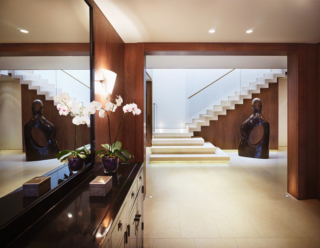 Vorraum mit Kommode vor Spiegel und Blick durch breiten Durchgang im eleganten Treppenraum auf moderne Skulptur
