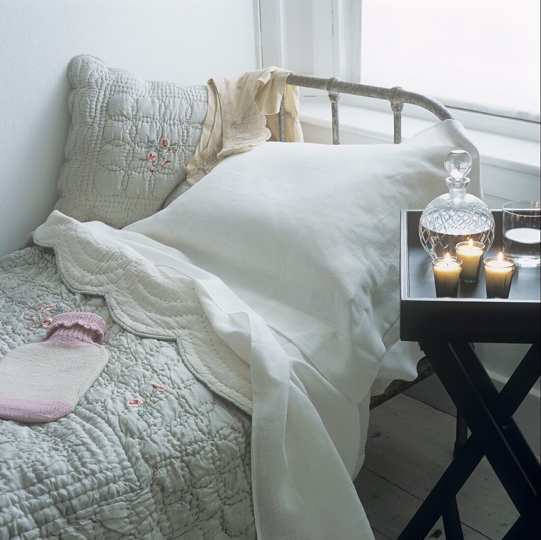 A bed with bedspread and hot water bottle and a folding table with candles and water