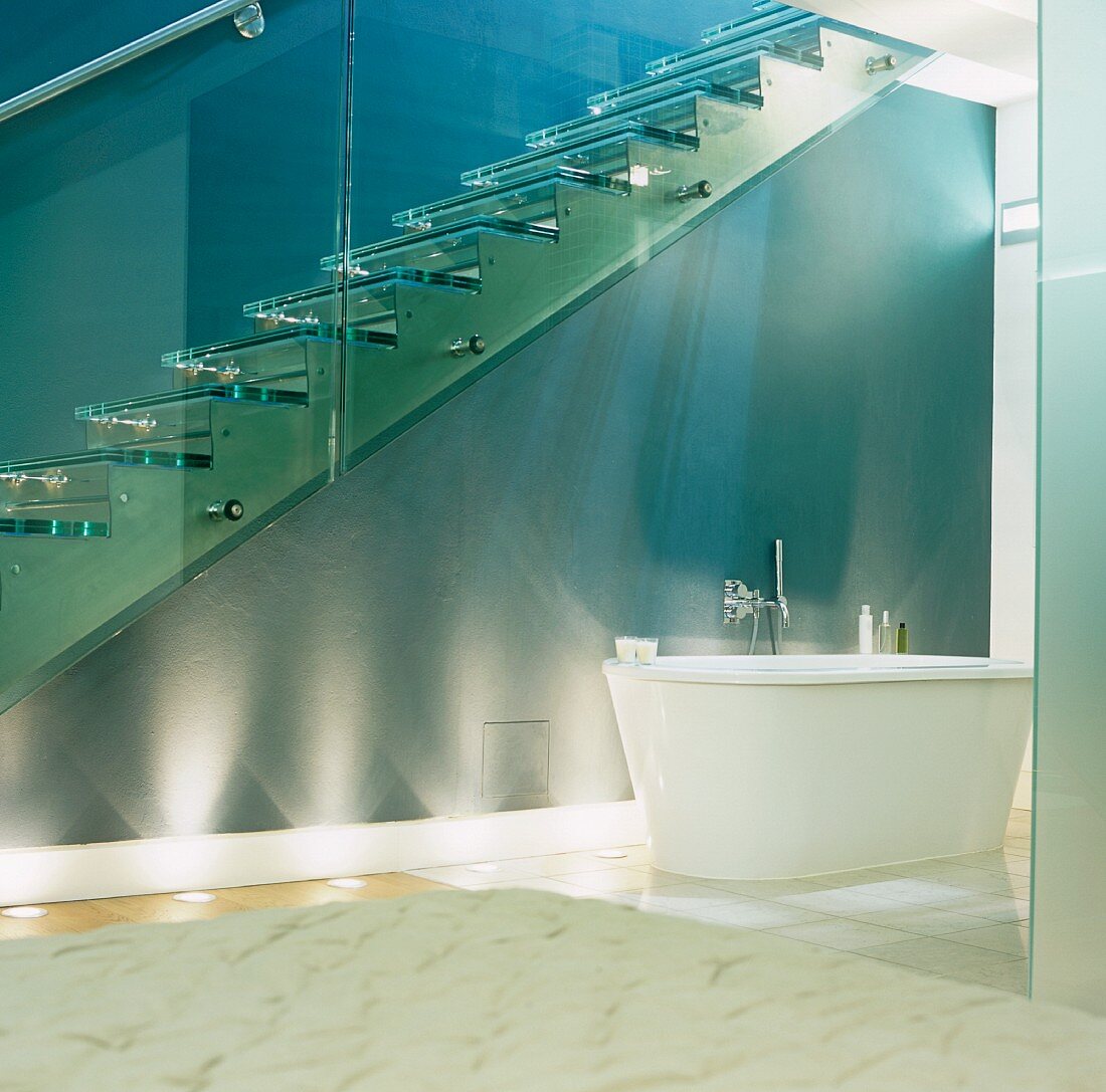 A free standing bathtub underneath a flight of glass stairs