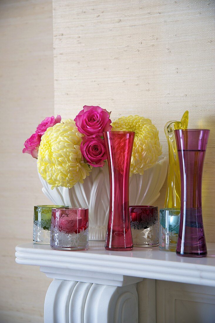 Farbige Glasvasen und Windlichter vor Blumenstrauss auf Kaminsims