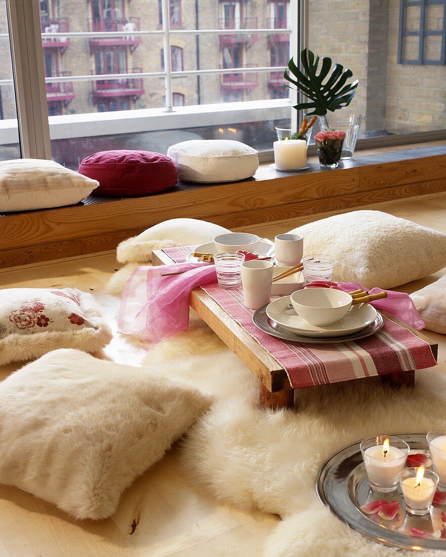 Bodenkissen aus weißem Fellbezug vor Bodentisch mit Gedeck und Fensterbank aus Holz
