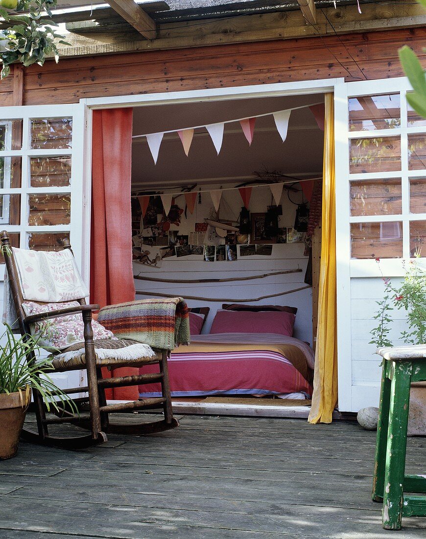Holzhaus mit Terrasse und Schaukelstuhl vor offener Tür mit Blick in Schlafraum