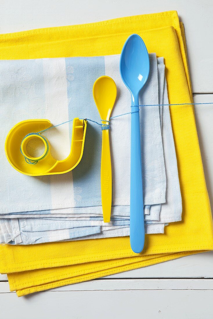 An arrangement of blue and yellow - a sticky tape dispenser and plastic spoons on a pile of tea towels