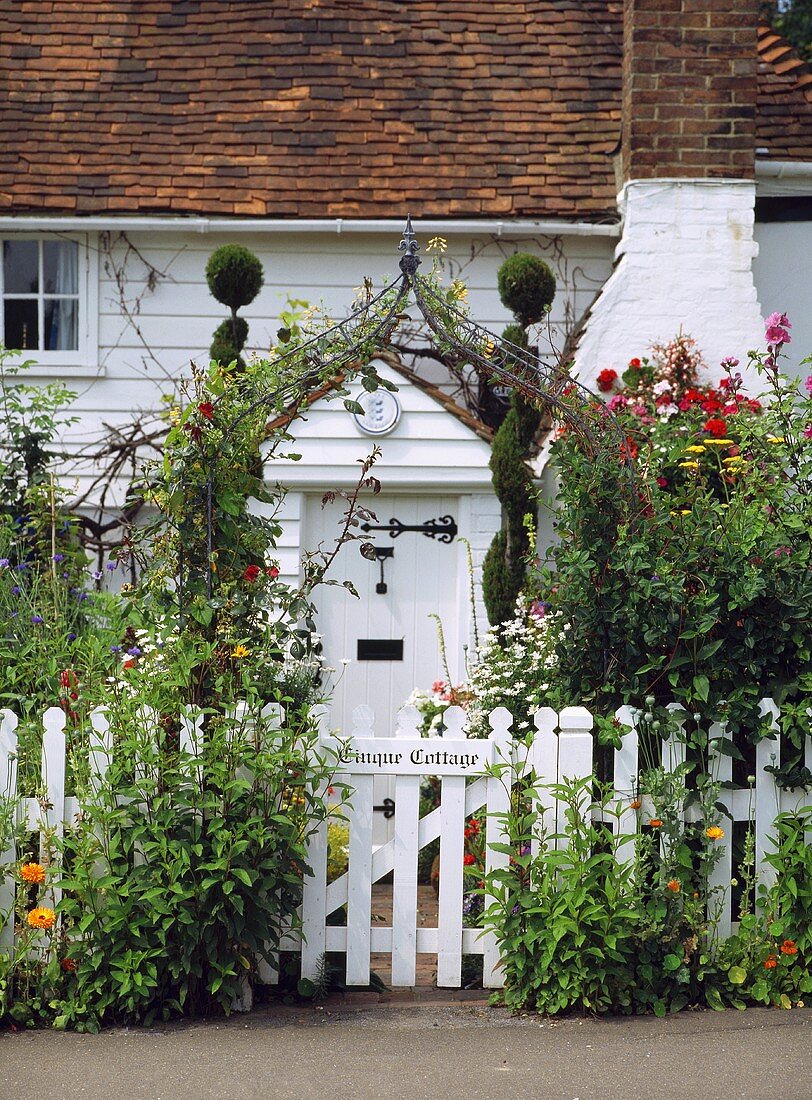 Begrünter weißer Gartenzaun mit Tür vor traditionellem Landhäuschen