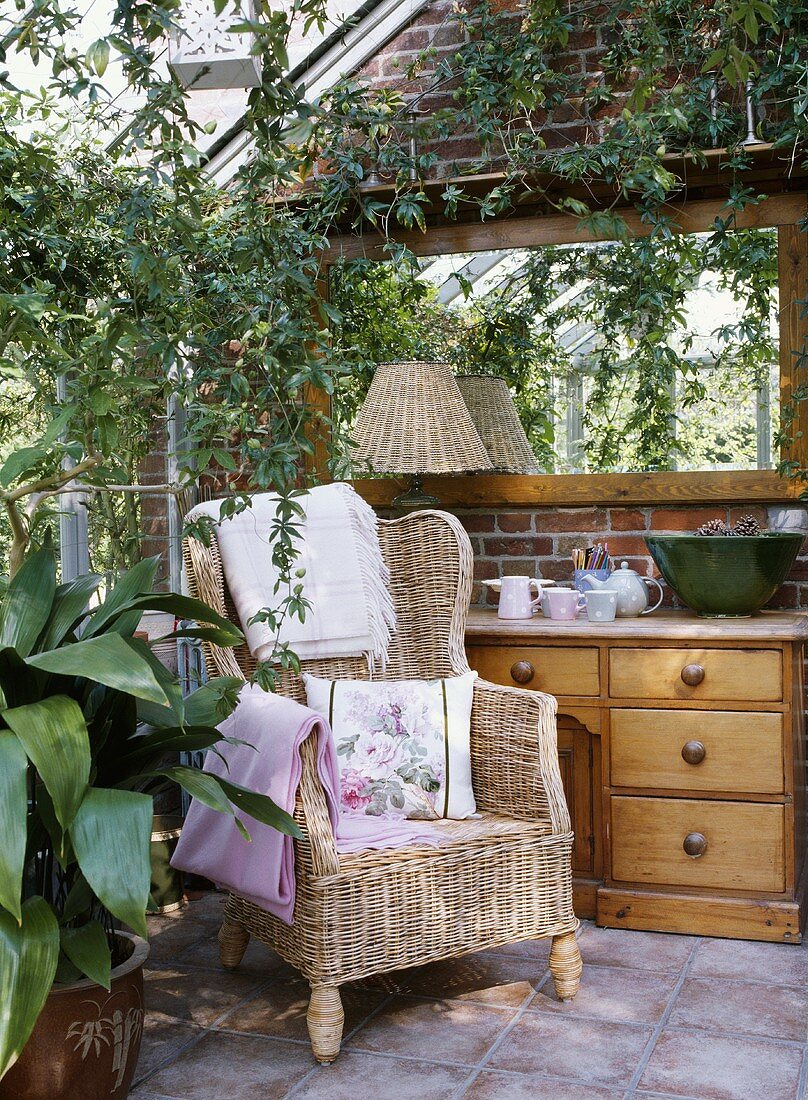 Wintergarten mit Korbsessel, einer Holzkommode mit Schubladen, einem darüber hängenden Spiegel und vielen Zimmerpflanzen