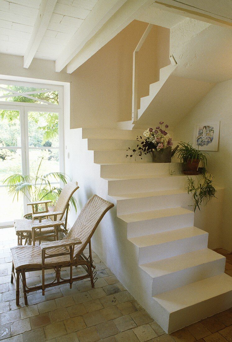 Rattan-Liegestühle unter weisser Steintreppe ohne Geländer in der Halle eines Landhauses