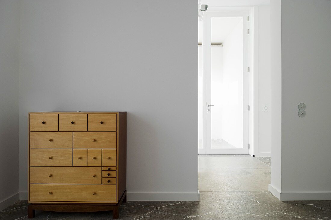 A simple wooden chest of drawers against a white wall and an open doorway with a view of a glass door