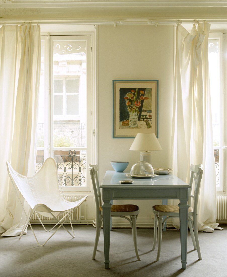 Französisches Esszimmer mit pastellblauem Tisch, weissen Bugholzstühlen und Butterfly-Stuhl vor lässig langen Vorhängen und Stillleben