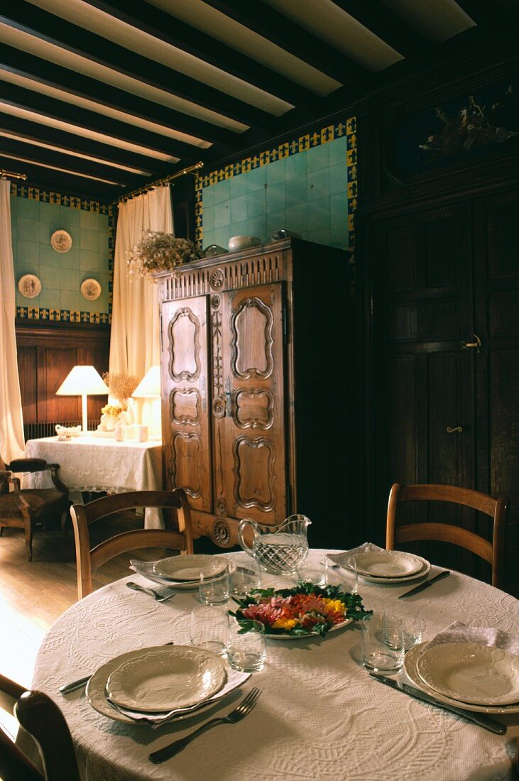 Gedecke auf weißem Tischtuch und geschnitzter Schrank unter Wandfliesen mit Bordüre in provenzalischem Muster