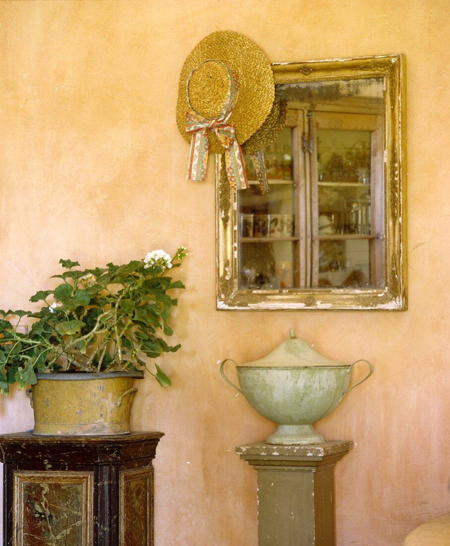 Antiker Spiegel mit Strohhut über Urne auf Sockel vor apricotfarbiger Wand