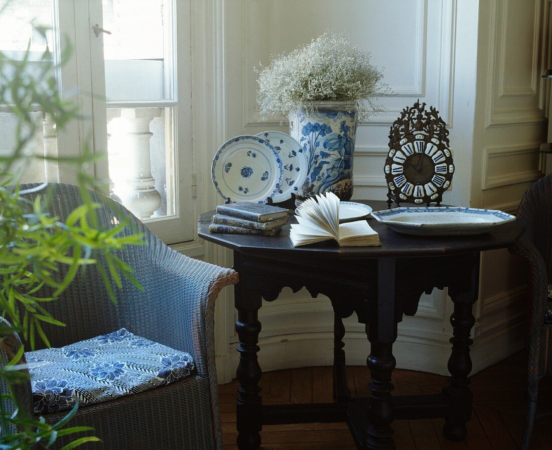 Englischer, blauer Korbsessel neben antikem Tisch mit blau-weißem, chinesischem Porzellan und Uhr in traditionellem Wohnzimmer