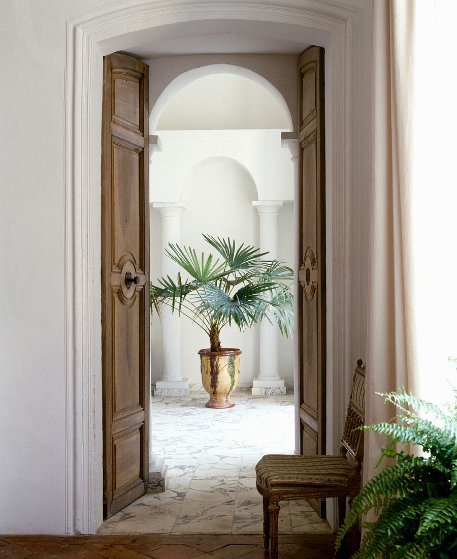 Blick durch grosse Kassetten-Doppeltüren auf Marmorboden mit Palme in glasiertem Keramiktopf vor weißem Säulenpaar