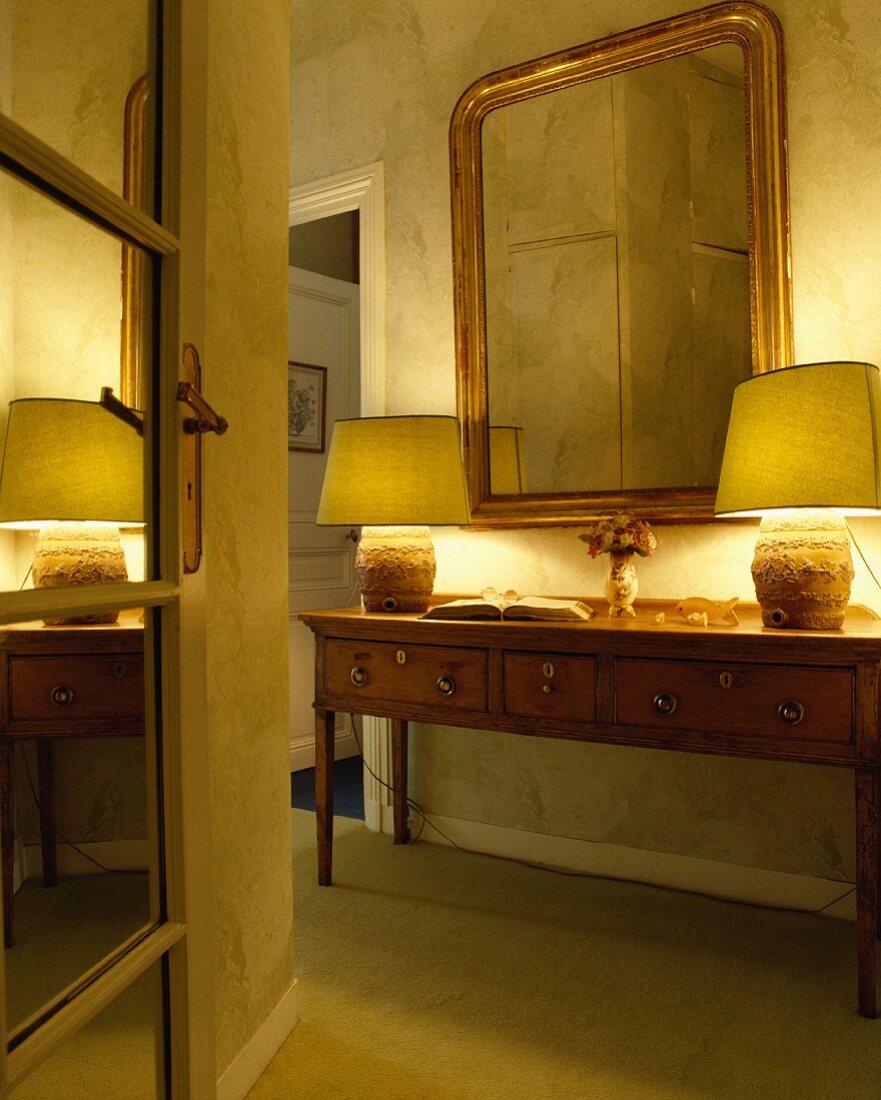 Blick durch offene Spiegeltür auf goldgerahmten Wandspiegel und brennende Lampen auf antiker Kiefern-Konsole